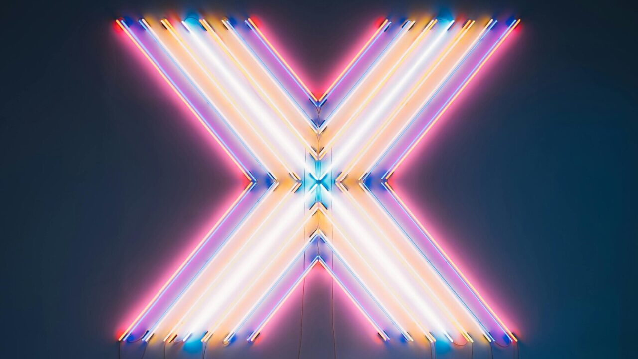 Vibrant neon X sign with colorful reflections on a dark floor background.