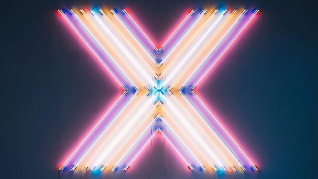 Vibrant neon X sign with colorful reflections on a dark floor background.